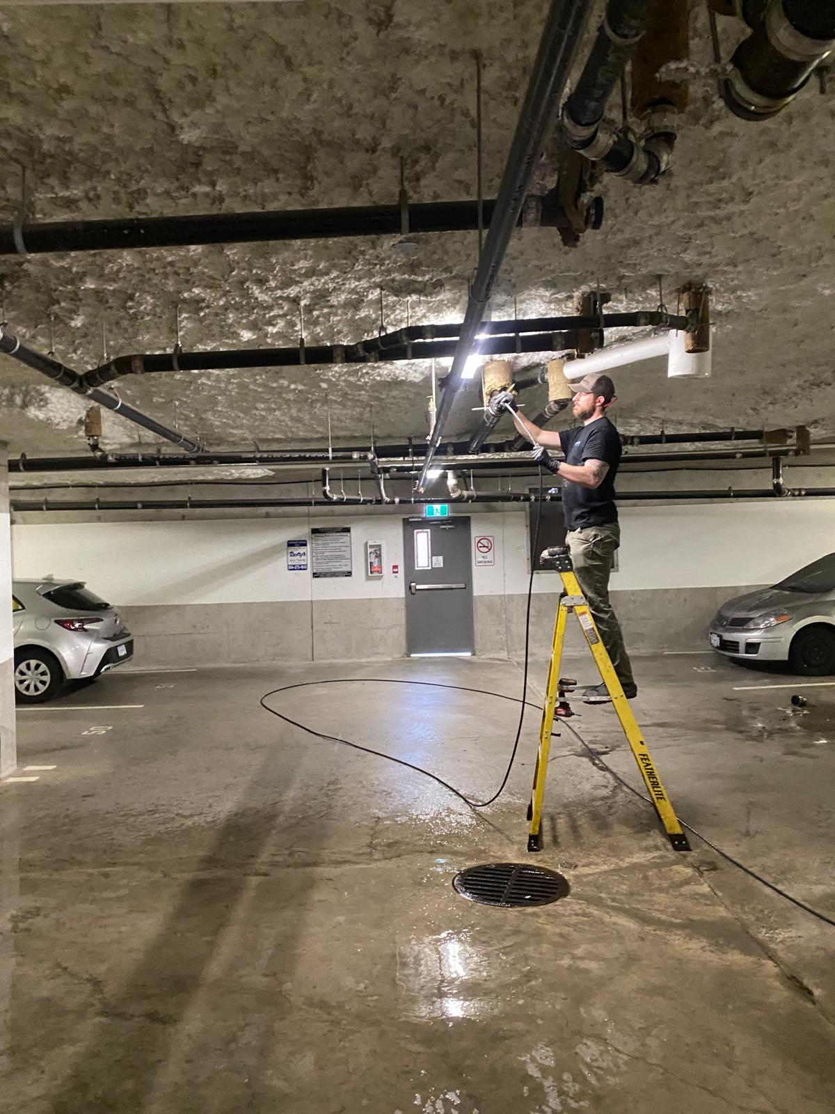 Technician on yellow ladder working on overhead drain pipe in parkade, commercial plumbing in Burnaby